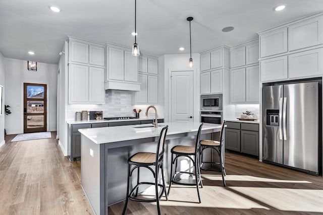 kitchen with appliances with stainless steel finishes, sink, pendant lighting, light hardwood / wood-style flooring, and an island with sink