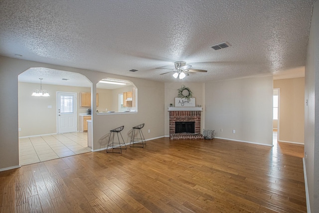 unfurnished living room featuring a textured ceiling, ceiling fan with notable chandelier, light hardwood / wood-style floors, and a brick fireplace