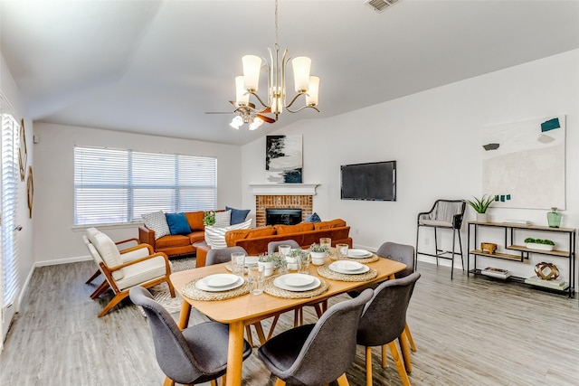 dining room with ceiling fan with notable chandelier, vaulted ceiling, light hardwood / wood-style flooring, and a brick fireplace