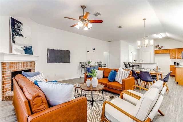 living room featuring ceiling fan with notable chandelier, light wood-type flooring, lofted ceiling, and a brick fireplace