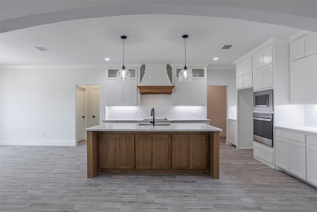 kitchen featuring premium range hood, a center island with sink, white cabinets, ornamental molding, and appliances with stainless steel finishes