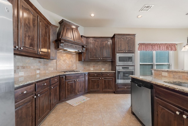 kitchen featuring light stone counters, dark brown cabinets, custom range hood, and appliances with stainless steel finishes