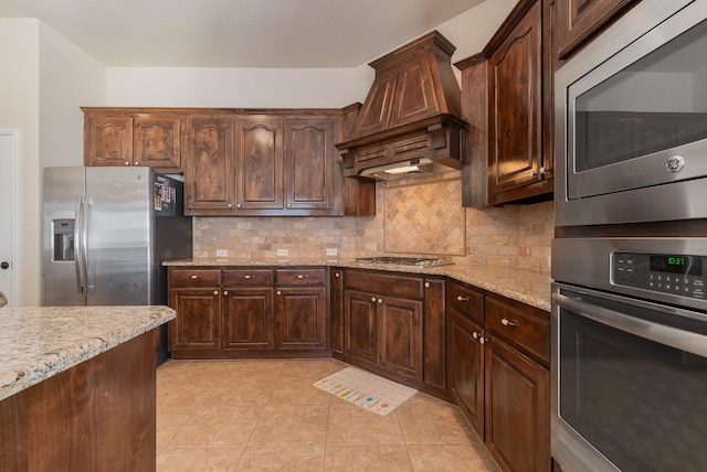 kitchen featuring light stone countertops, backsplash, custom range hood, dark brown cabinets, and stainless steel appliances