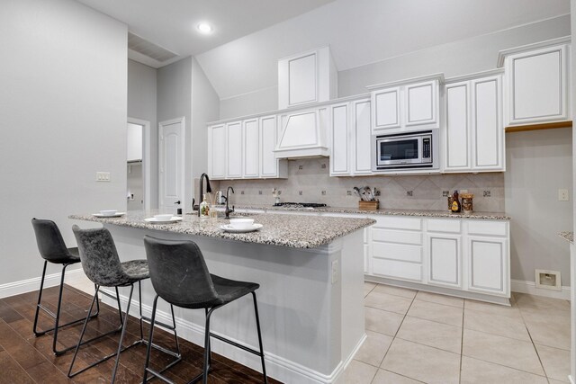 kitchen with premium range hood, stainless steel microwave, an island with sink, white cabinets, and decorative backsplash