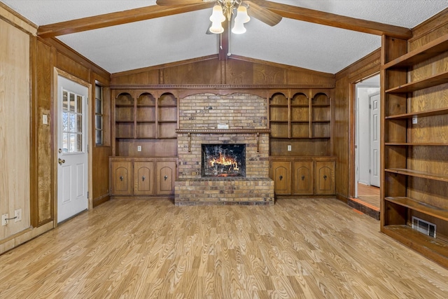 unfurnished living room with wood walls, lofted ceiling with beams, a textured ceiling, a brick fireplace, and light hardwood / wood-style flooring