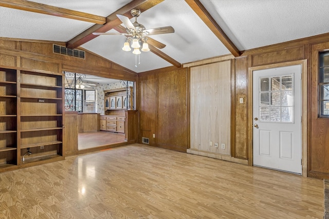 unfurnished living room with ceiling fan, wooden walls, lofted ceiling with beams, a textured ceiling, and light wood-type flooring