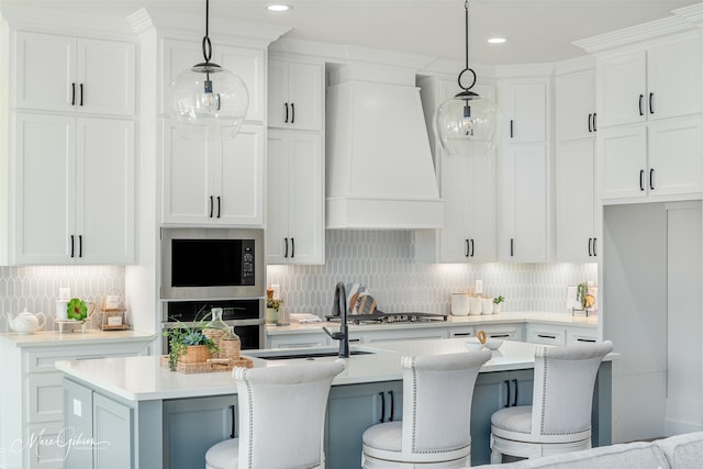 kitchen with white cabinetry, hanging light fixtures, stainless steel appliances, premium range hood, and a kitchen island with sink