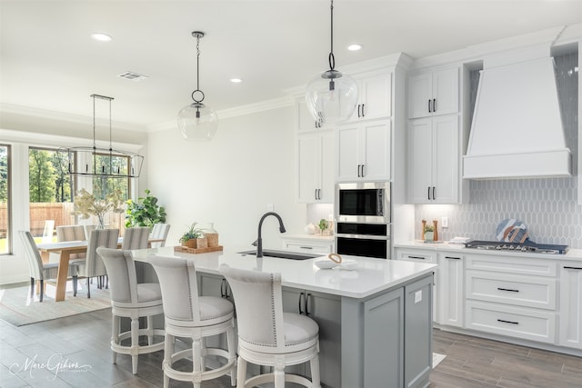 kitchen featuring custom exhaust hood, sink, pendant lighting, a center island with sink, and white cabinetry