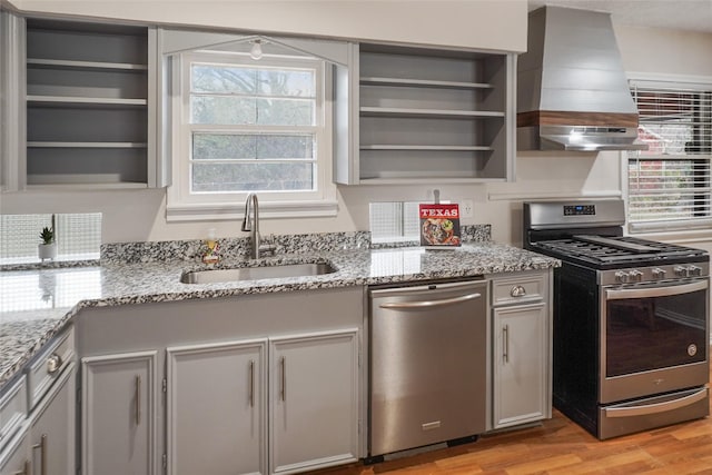 kitchen with gray cabinetry, wall chimney range hood, sink, light stone counters, and stainless steel appliances