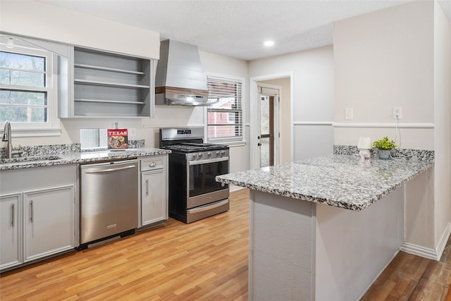 kitchen featuring sink, wall chimney exhaust hood, light stone countertops, appliances with stainless steel finishes, and kitchen peninsula