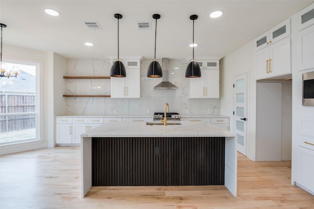 kitchen with visible vents, glass insert cabinets, an island with sink, light stone countertops, and wall chimney exhaust hood