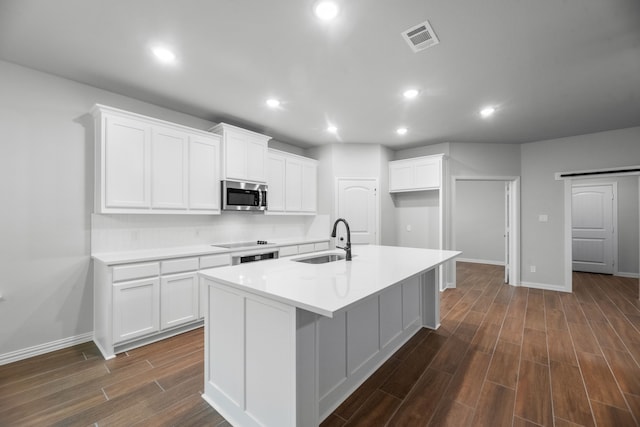 kitchen with black electric stovetop, stainless steel microwave, a sink, dark wood-type flooring, and a center island with sink