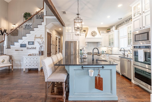 kitchen with appliances with stainless steel finishes, custom range hood, sink, a center island with sink, and white cabinets