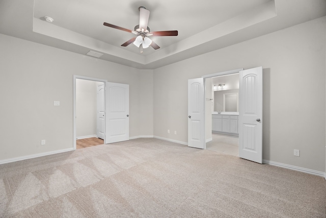 unfurnished bedroom featuring light colored carpet, ensuite bathroom, a tray ceiling, baseboards, and a ceiling fan
