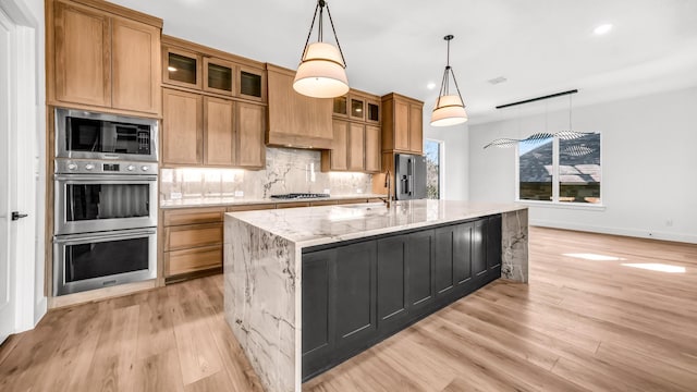 kitchen featuring a center island with sink, decorative backsplash, appliances with stainless steel finishes, light stone countertops, and a sink