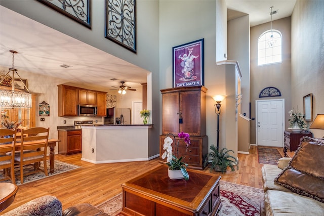 living area featuring baseboards, visible vents, a towering ceiling, light wood-style floors, and ceiling fan with notable chandelier