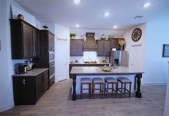 kitchen featuring stainless steel appliances, a kitchen bar, a center island with sink, custom range hood, and hardwood / wood-style flooring