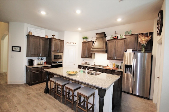 kitchen with premium range hood, a center island with sink, sink, appliances with stainless steel finishes, and a kitchen bar