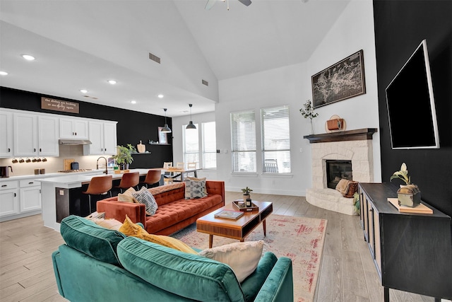 living room featuring ceiling fan, high vaulted ceiling, a stone fireplace, and light hardwood / wood-style floors