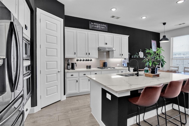 kitchen featuring light stone counters, stainless steel appliances, a kitchen island with sink, sink, and hanging light fixtures