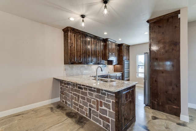 kitchen featuring kitchen peninsula, backsplash, light stone counters, dark brown cabinets, and sink