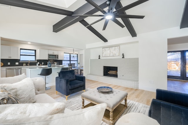 living room with lofted ceiling with beams, light hardwood / wood-style floors, ceiling fan with notable chandelier, and a brick fireplace