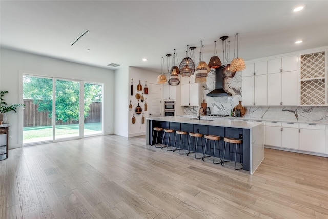 kitchen featuring wall chimney range hood, decorative light fixtures, white cabinetry, and an island with sink