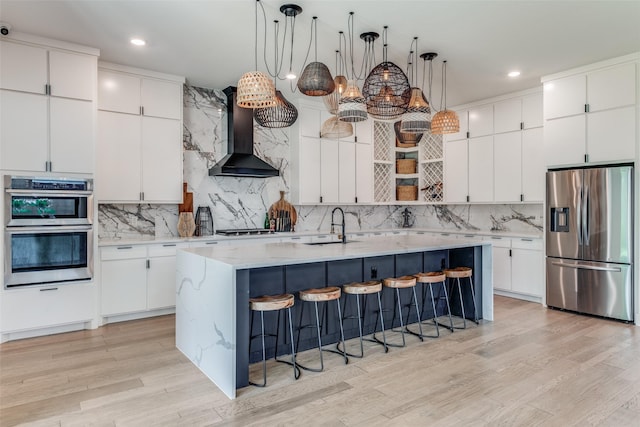 kitchen featuring stainless steel appliances, a kitchen island with sink, wall chimney range hood, decorative light fixtures, and white cabinets