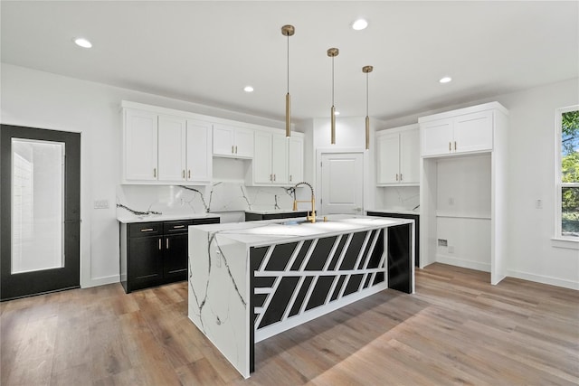 kitchen with pendant lighting, a kitchen island with sink, backsplash, white cabinetry, and light hardwood / wood-style floors