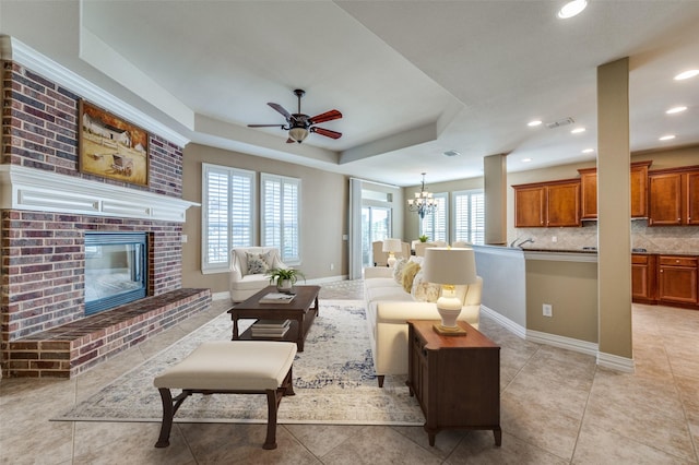 living room featuring ceiling fan with notable chandelier, light tile patterned flooring, a fireplace, and a tray ceiling