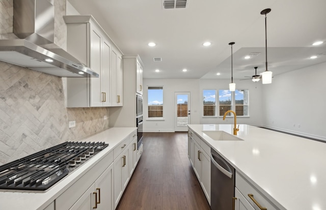 kitchen featuring hanging light fixtures, stainless steel appliances, white cabinets, wall chimney range hood, and sink