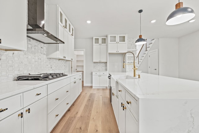 kitchen with white cabinetry, wall chimney exhaust hood, hanging light fixtures, stainless steel gas stovetop, and a kitchen island with sink