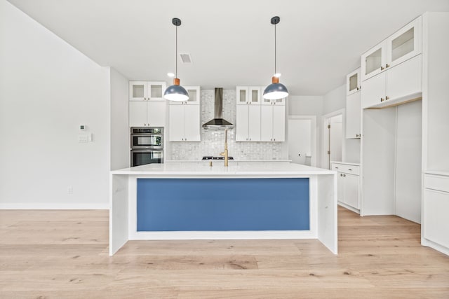 kitchen with appliances with stainless steel finishes, wall chimney exhaust hood, pendant lighting, a center island with sink, and white cabinets