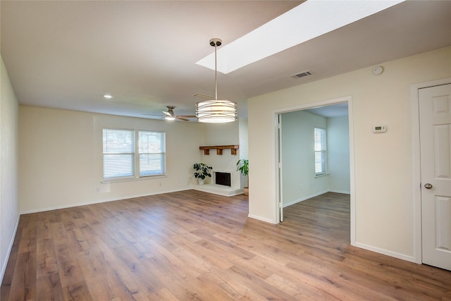 unfurnished living room featuring ceiling fan, wood-type flooring, a skylight, and a brick fireplace