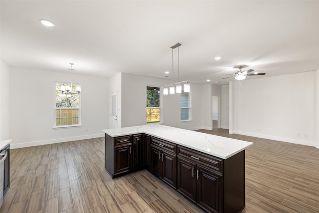 kitchen with light stone countertops, a center island, pendant lighting, dark brown cabinets, and ceiling fan with notable chandelier