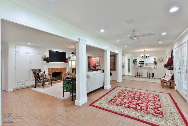 living room featuring ornate columns, ornamental molding, ceiling fan with notable chandelier, and a brick fireplace