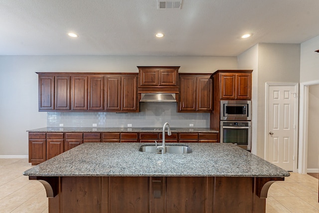 kitchen with sink, a center island with sink, stainless steel appliances, light stone countertops, and backsplash