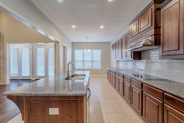 kitchen featuring sink, light stone counters, pendant lighting, black electric stovetop, and a center island with sink