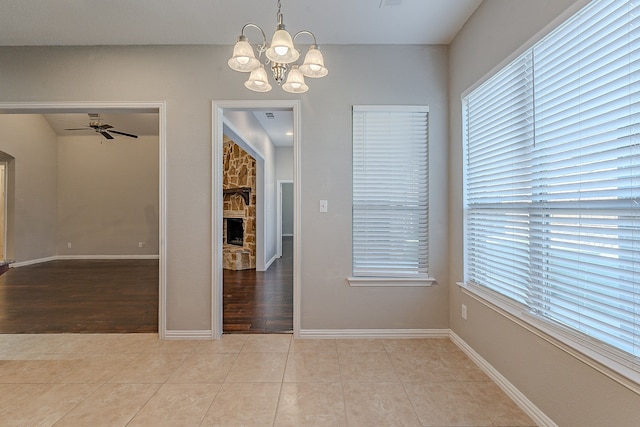 unfurnished dining area with a stone fireplace, light tile patterned floors, and ceiling fan with notable chandelier