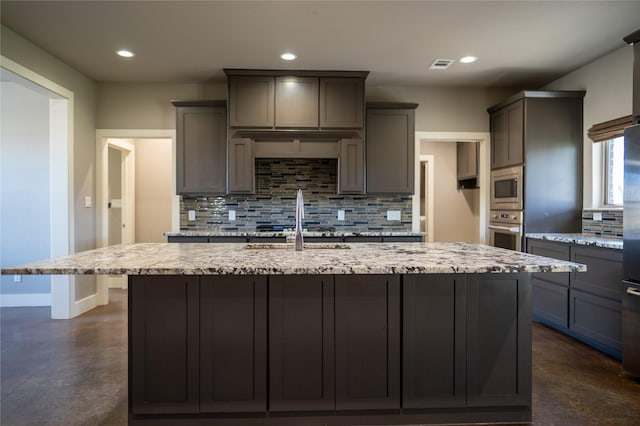 kitchen with backsplash, light stone countertops, an island with sink, and stainless steel appliances