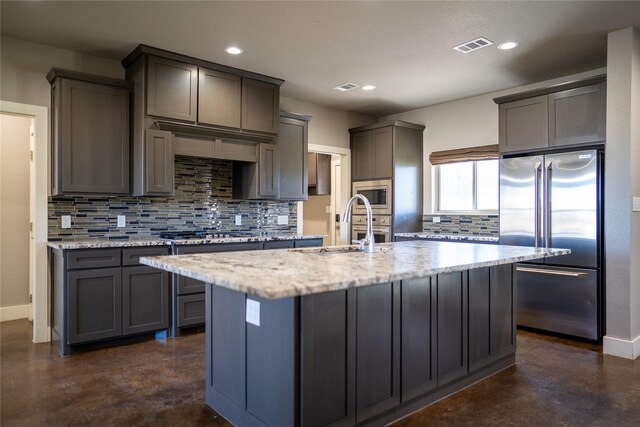 kitchen featuring decorative backsplash, light stone counters, an island with sink, and appliances with stainless steel finishes