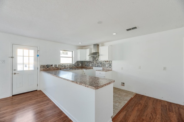 kitchen featuring wall chimney range hood, light hardwood / wood-style flooring, white cabinetry, tasteful backsplash, and kitchen peninsula