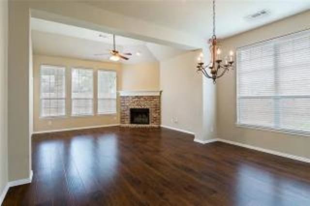 unfurnished living room with a brick fireplace, plenty of natural light, ceiling fan with notable chandelier, and dark hardwood / wood-style floors