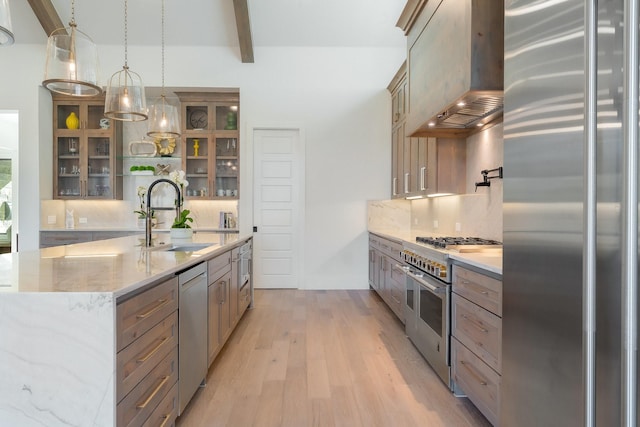 kitchen with sink, appliances with stainless steel finishes, a kitchen island with sink, decorative light fixtures, and wall chimney exhaust hood