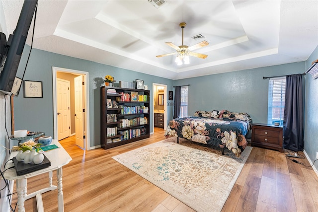 bedroom with ceiling fan, ensuite bath, a tray ceiling, and hardwood / wood-style floors