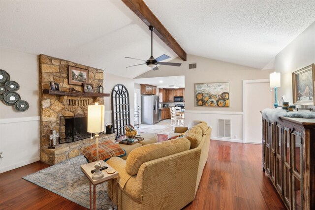 living room featuring hardwood / wood-style floors, a stone fireplace, vaulted ceiling with beams, ceiling fan, and a textured ceiling