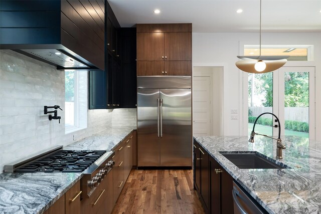 kitchen with light stone countertops, stainless steel appliances, sink, wall chimney range hood, and hanging light fixtures