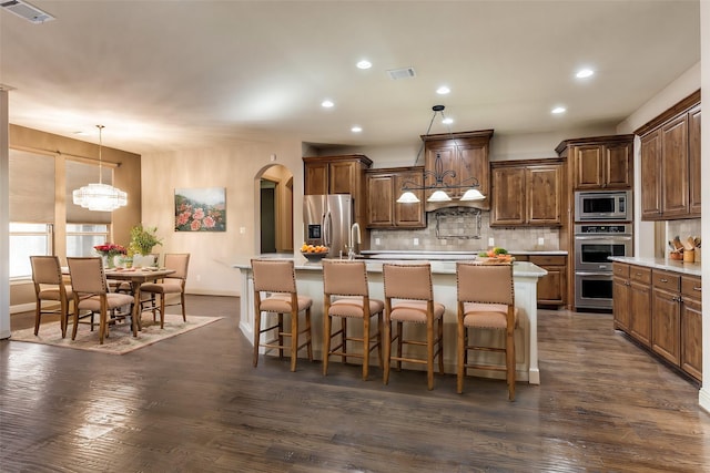 kitchen with decorative backsplash, stainless steel appliances, dark wood-type flooring, decorative light fixtures, and an island with sink