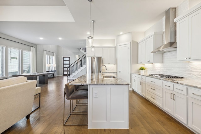 kitchen with white cabinetry, sink, wall chimney exhaust hood, stainless steel appliances, and a center island with sink