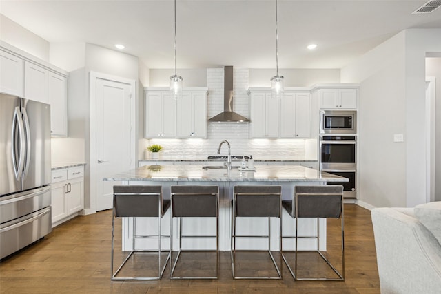 kitchen with appliances with stainless steel finishes, pendant lighting, a kitchen island with sink, and wall chimney exhaust hood